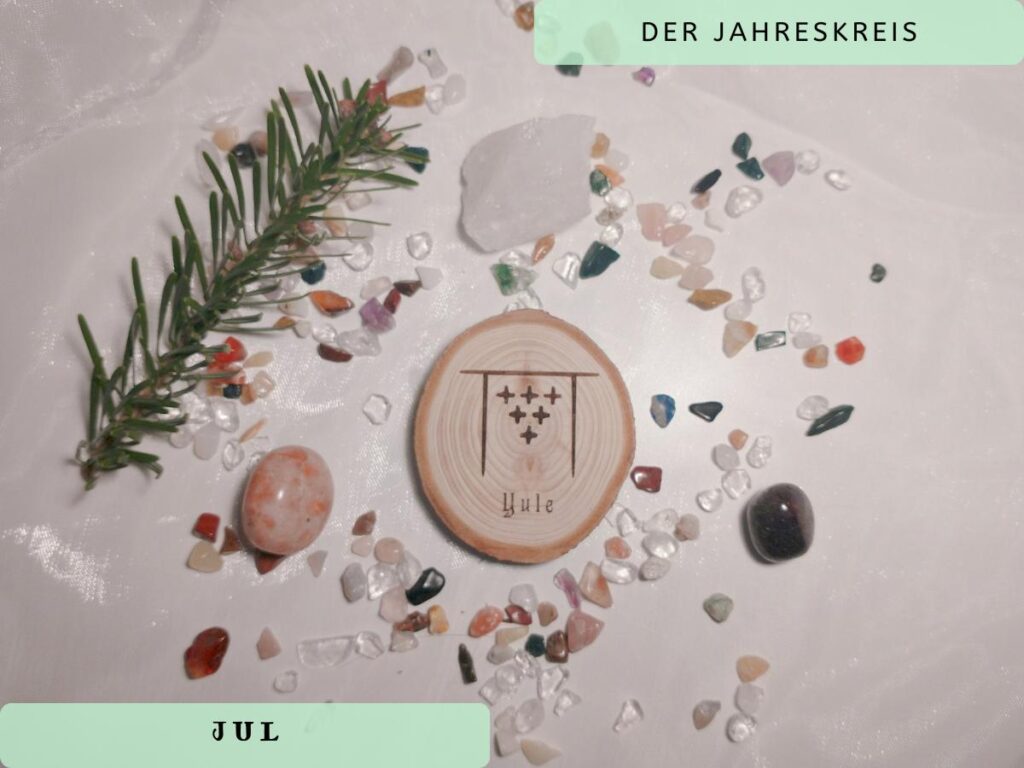 Das Bild zeigt die Rune von Jul Yule eingeprägt auf einer Baumscheibe und als Symbole für Jul einen Tannenzweis und die Heilsteine Bergkristall, Granat und Sonnenstein. Alles liegt auf einem weißen Chiffontuch mit kleinen Edelsteinchips.